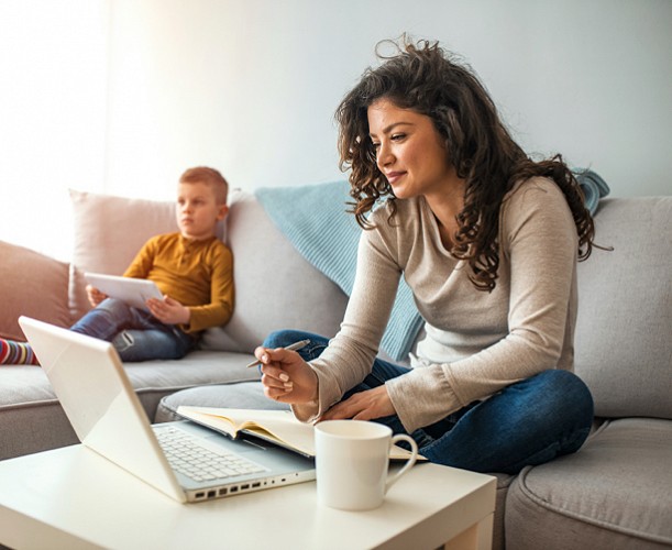 Person sitting on couch working on laptop with child in background.